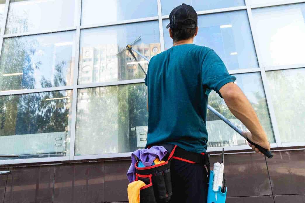 A window cleaner wearing a cap and carrying cleaning supplies uses a long pole to clean the large glass windows of a commercial building.
